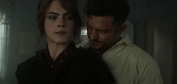 A screenshot from the new trailer of season 2 of Carnival Row shows actors Cara Delevingne and Orlando Bloom standing close together in a large room. Cara Delevingne's character Vignette Stonemoss is wearing a black dress and is looking worried and Orlando Bloom's character Rycroft "Philo" Philostrate is wearing a white shirt as he stands close behind Vignerre