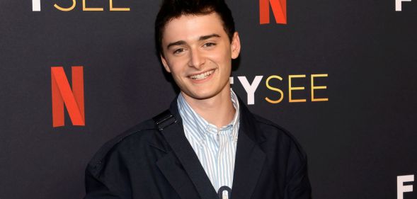 Noah Schnapp smiles while wearing a light coloured shirt and dark coloured jacket while standing in front of a background with the Netflix logo on it