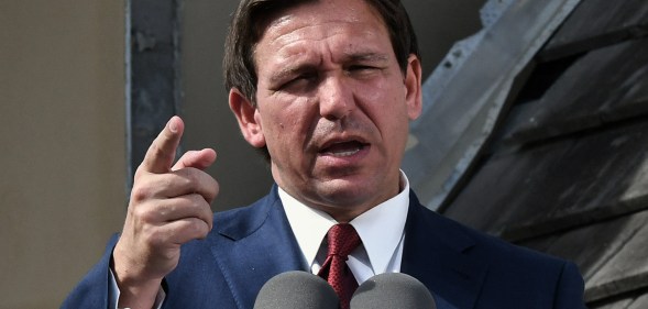 Florida governor Ron DeSantis wearing a navy suit, white shirt and red tie, stands in front of a microphone at a press conference