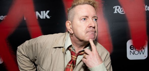 John Lydon wearing a beige jacket and checkered tie looking to the right with his finger on his chin.