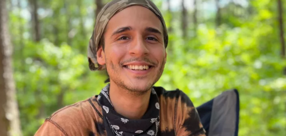 A photo of queer environmental activist Manuel “Tortuguita” Terán wearing a light orange t-shirt and smiling, with a forest in the background