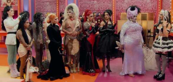 A screenshot from RuPaul’s Drag Race season 15 shows the drag queen contestants standing next to each other in one of the series' sets that has a orange and pink floor with pink-pattern walls and lights in the background