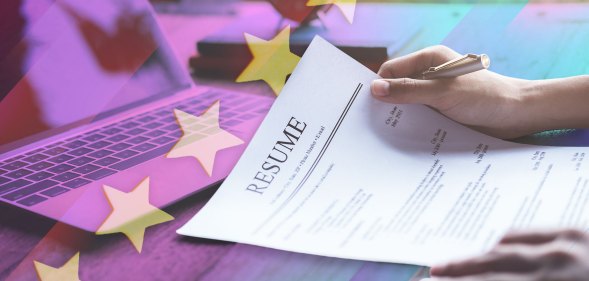 A pair of hands are holding a CV on a desk with a computer in the background.