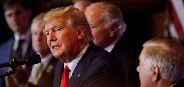 A photo shows Donald Trump wearing a navy suit, whit shirt and red tie speaking to an audience during a campaign speech.