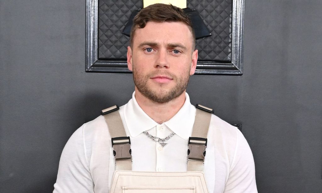 Gus Kenworthy at the 2023 Grammy Awards wearing a white shirt and beige and grey dungarees