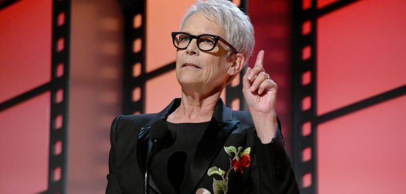 A photo of actor Jamie Lee Curtis wearing glasses and a black dress on stage at the AARP awards in LA.