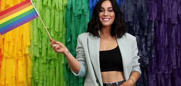 Sophie Cachia wearing a black top and grey blazer stands with one hand in her pocket and the other holding a pride flag.