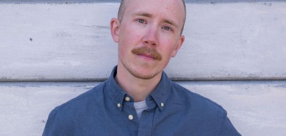 Trans activist and journalist Freddy McConnell wears a blue shirt as he stands in front of panelling