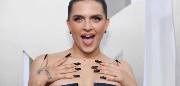 Mae Muller wears a black dress at the MTV 2022 VMAs, looking off camera, hands on her shoulders, and with her mouth open.