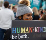 A person holds a sign reading 'Human-kind, be both' written in rainbow script at an LGBTQ+ rights protest