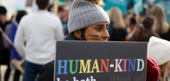 A person holds a sign reading 'Human-kind, be both' written in rainbow script at an LGBTQ+ rights protest