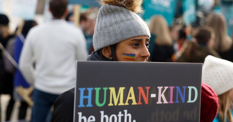 A person holds a sign reading 'Human-kind, be both' written in rainbow script at an LGBTQ+ rights protest