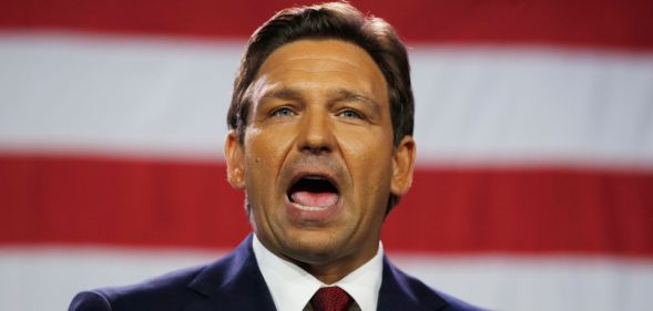 Florida governor Ron DeSantis wears a suit and tie as he speaks with his mouth wide open. The red and white stripes of the US flag can be seen in the background