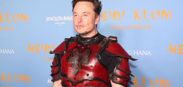 Twitter boss Elon Musk wears a red and black armour like outfit as he stands in front of a light blue and yellow background