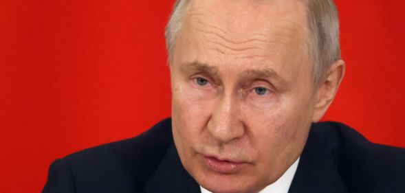 Russia president Vladimir Putin, who has a history of attacking LGBTQ+ people, wears a suit and tie as he sits in front of a red background