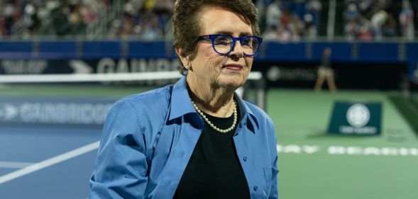 Tennis legend Billie Jean King wears a dark shirt and blue jacket as she stands on a tennis court