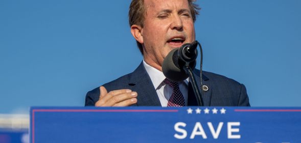 Ken Paxton speaking to a crowd, with a sign reading "save America" below him.