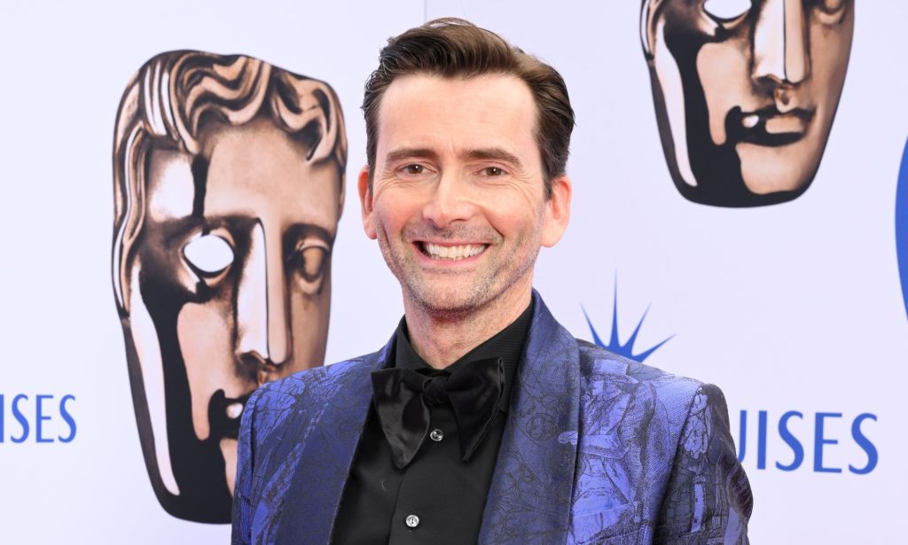 David Tennant wears a blue suit and black shirt while smiling at the camera.