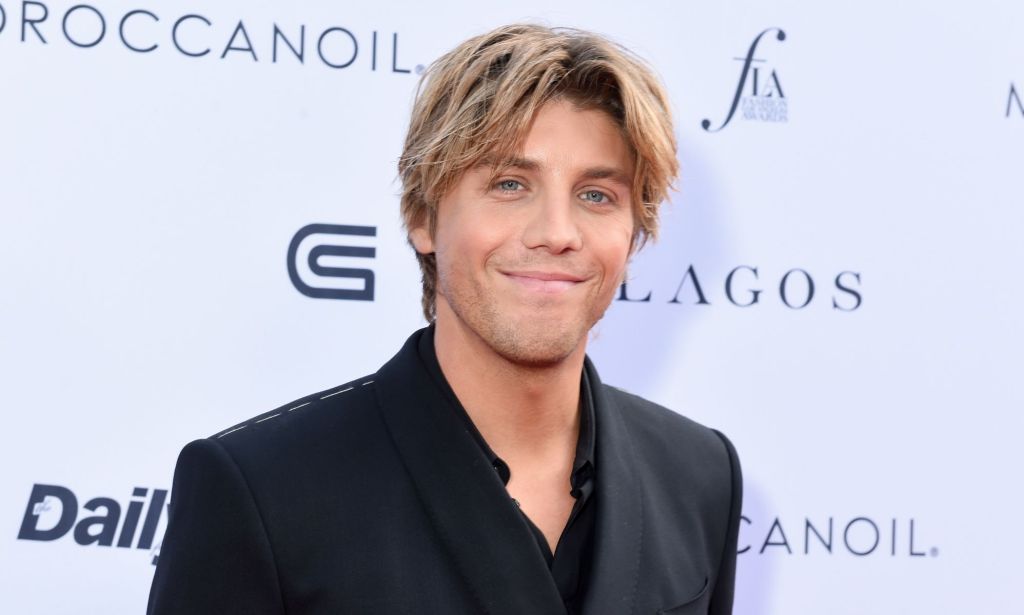 Lukas Gage in a black suit and shirt with parted blonde hair, smiling at the camera.