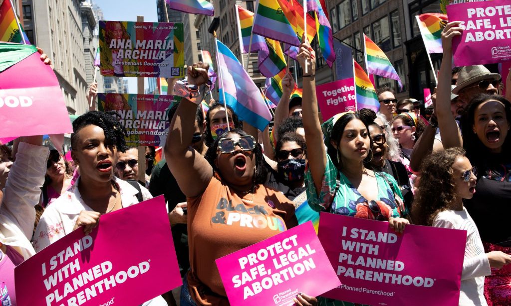 A crowd of people hold up signs in favour of reproductive rights and denouncing the Supreme Court for overturning Roe v Wade while holding LGBTQ+ and trans Pride flags