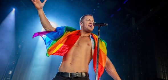 Imagine Dragons frontman Dan Reynolds performs shirtless on stage with an LGBTQ+ flag wrapped around his shoulders.