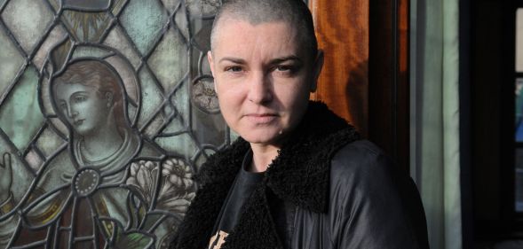 Sinead O'Connor wears a black top and jacket while standing in front of a front door.