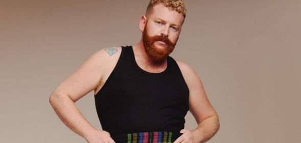 Non-binary, trans singer Tom Rasmussen wears a black vest and tartan shorts against a beige background.