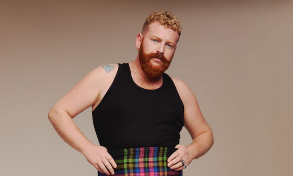 Non-binary, trans singer Tom Rasmussen wears a black vest and tartan shorts against a beige background.