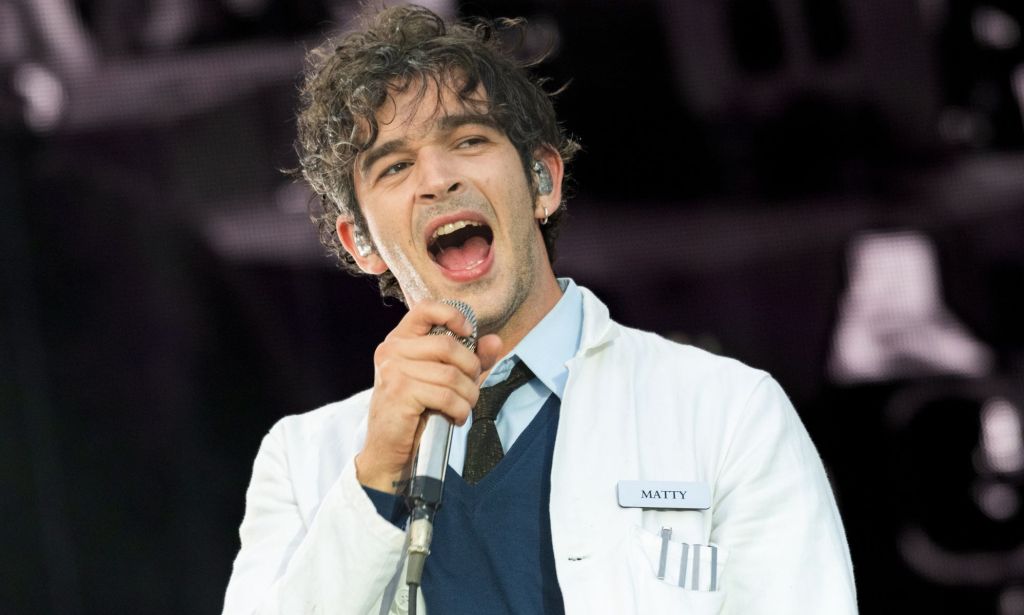 Matty Healy of The 1975 performing on stage wearing a lab coat, a sweater and a shirt and tie.