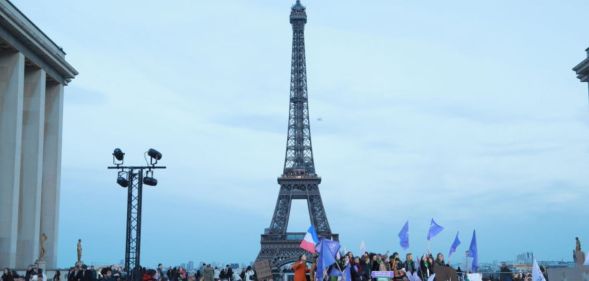 This is an image of the Eifel Tower in Paris, France. It is an overcast day. There is a large group of people in the foreground and we can see French flags flying.