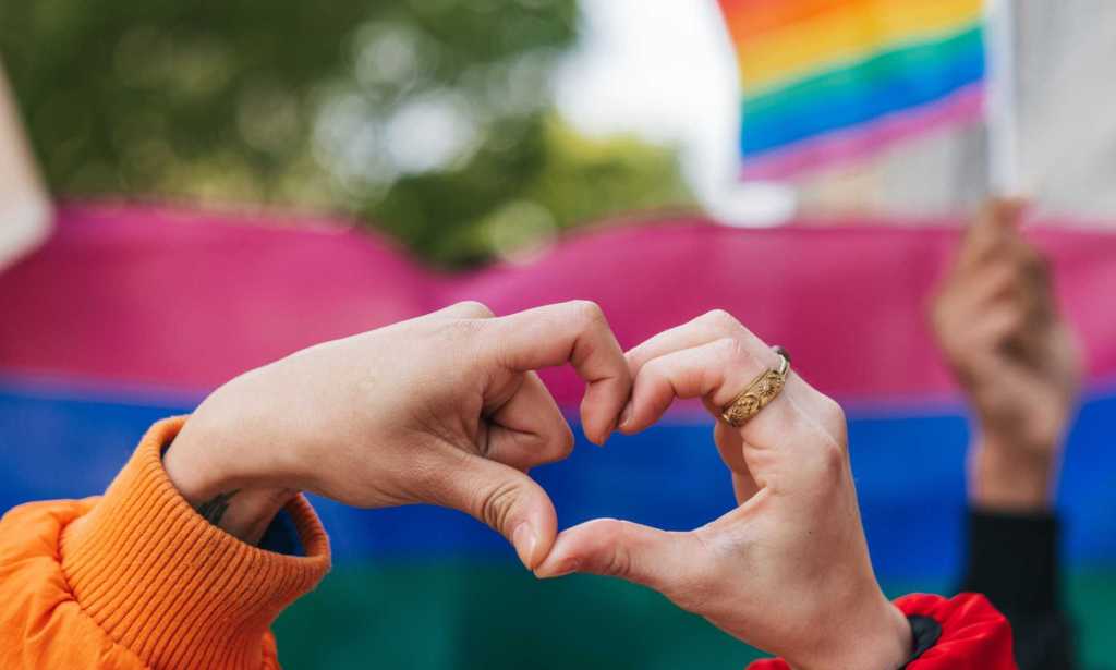 Genderqueer and non-binary friends making a heart shape against a rainbow flag celebrating love during a Pride Parade.