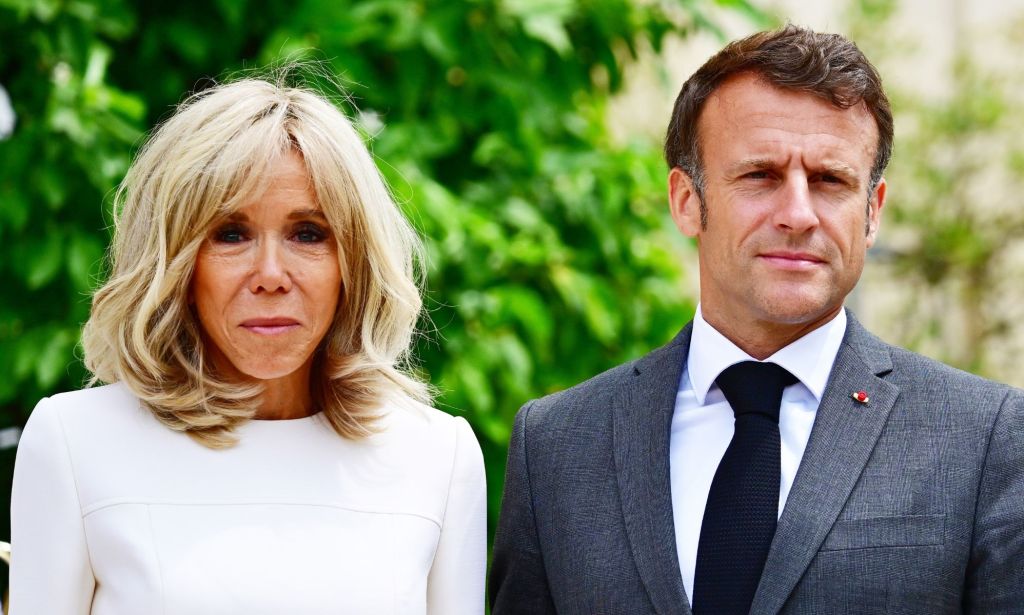 French president Emmanuel Macron wears a suit and tie as he stands beside his wife Brigitte, who is wearing white