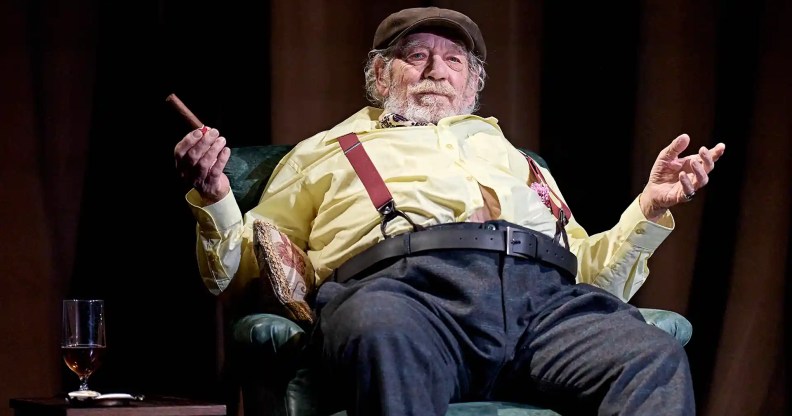 Ian McKellen in Player Kings, sitting on a chair, his chest and stomach fatter than usual, wearing a flat cap, yellow shirt and blue trousers, with red braces