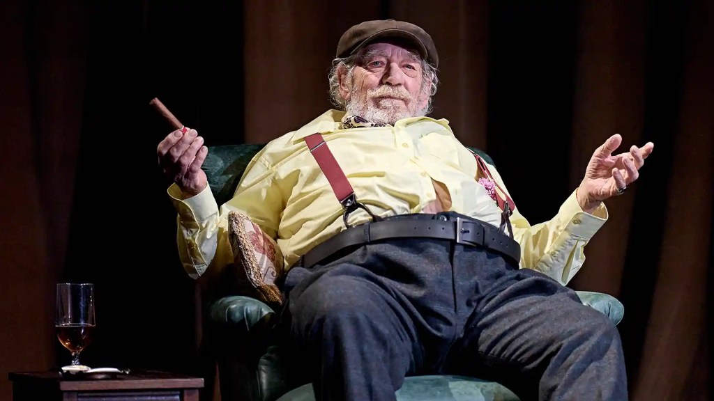 Ian McKellen in Player Kings, sitting on a chair, his chest and stomach fatter than usual, wearing a flat cap, yellow shirt and blue trousers, with red braces