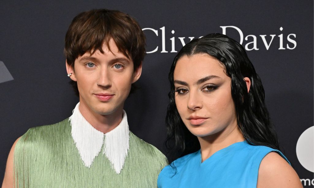 Troye Sivan in a frilly green top with a white collar standing next to Charli XCX who is wearing a blue armless dress. They are stood against a black background.