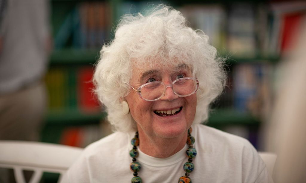 Trans author Jan Morris wears a white shirt and large necklace as she smiles towards someone off camera
