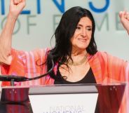Trans academic and activist Sandy Stone wears a dark shirt and pink top as she holds up her hands while being inducted into the US National Women's Hall of Fame