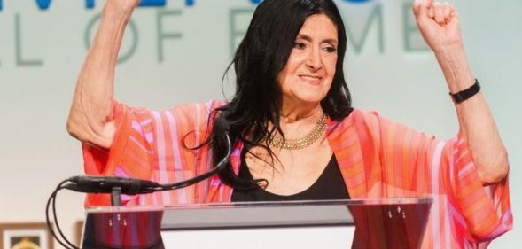 Trans academic and activist Sandy Stone wears a dark shirt and pink top as she holds up her hands while being inducted into the US National Women's Hall of Fame