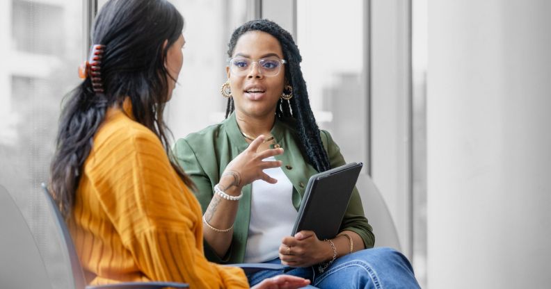 This is an image of two women talking. In the background there is a Black woman, wearing a green blazer, white tshirt and jeans. She is talking to someone wearing a yellow dress with long dark hair.