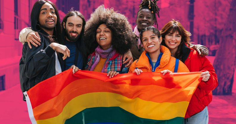 This is an image of a group of young and diverse LGBTQ+ people. They are posing for the camera and are holding a pride flag.
