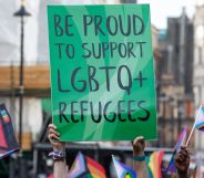 This is an image of a sign in black text on a green board. The text reads "Be Proud to support LGBTQ+ refugees". THere are various pride flags in hand