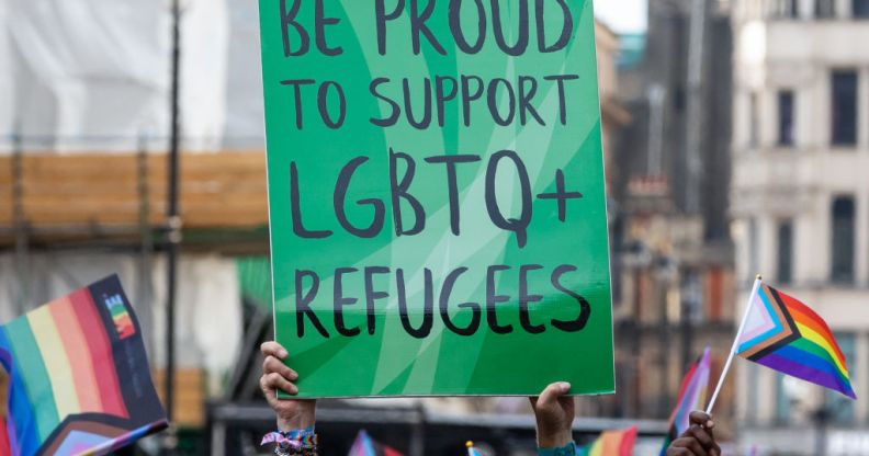 This is an image of a sign in black text on a green board. The text reads "Be Proud to support LGBTQ+ refugees". THere are various pride flags in hand