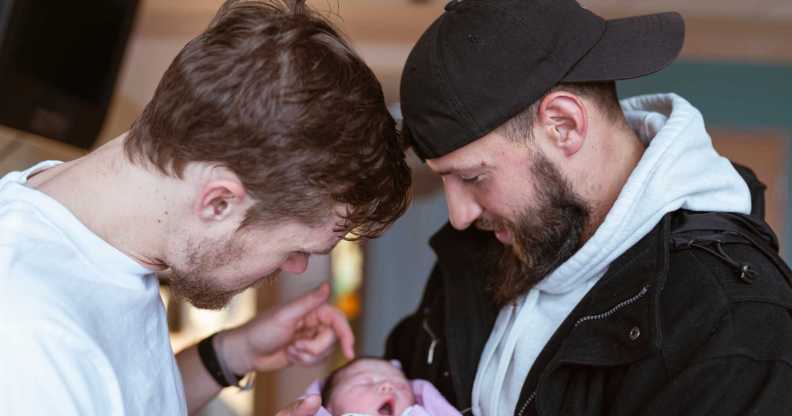 Two caucasian men in their thirties are affectionally holding and looking at a newborn baby lovingly.