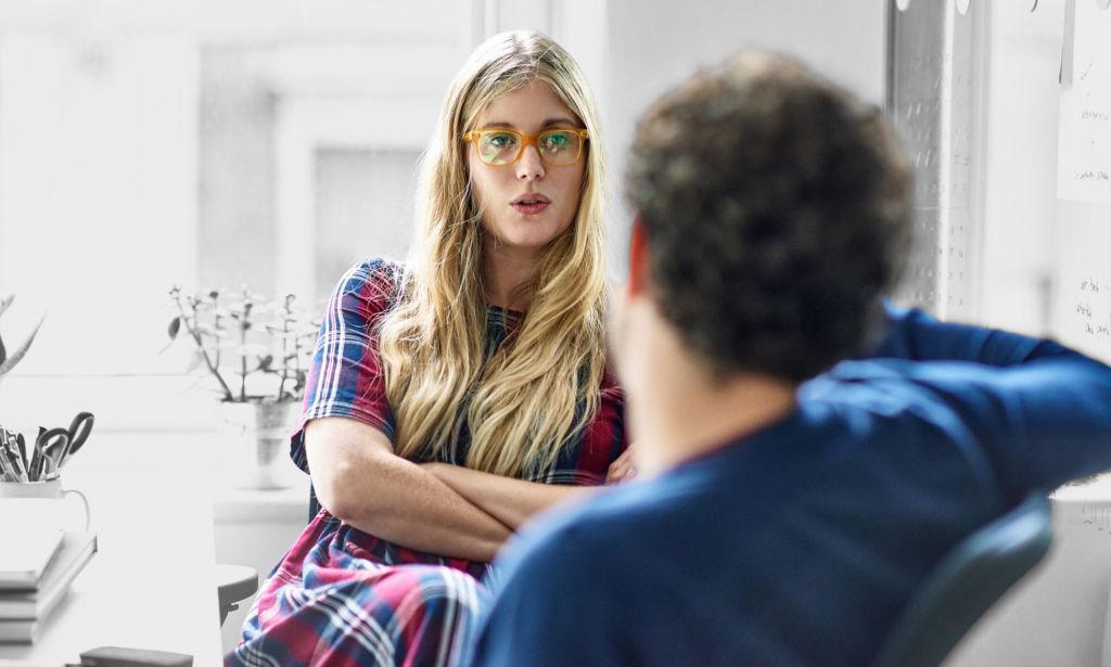 This is an image of two people having a tense conversation. In the background there is a woman with blonde hair, her arms crossed and wearing eyeglasses. She is speaking to someone in the foreground wearing a blue jumper with short curly hair. 