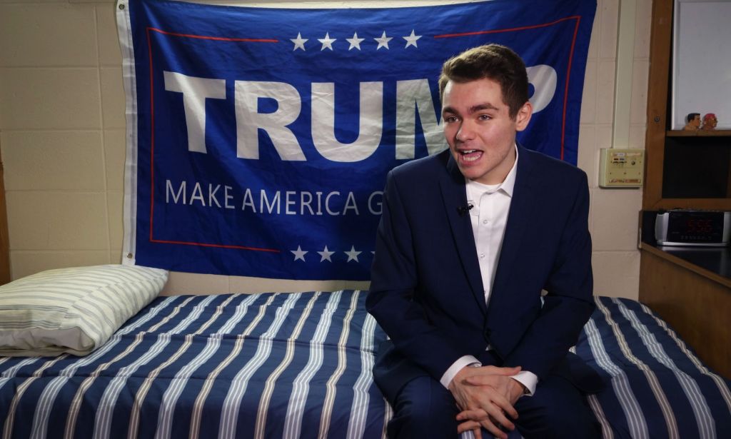 Nick Fuentes sitting on a bed in front of a Trump flag.