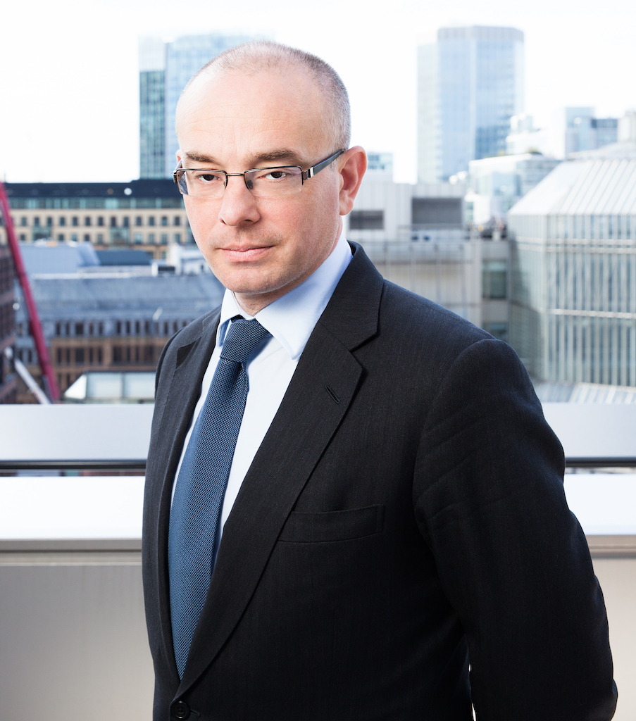 This is an image of a white man standing outside with the London skyline behind him. He is is bald and wearing glasses. He is wearing a dark suit with a dark bluish-green tie.
