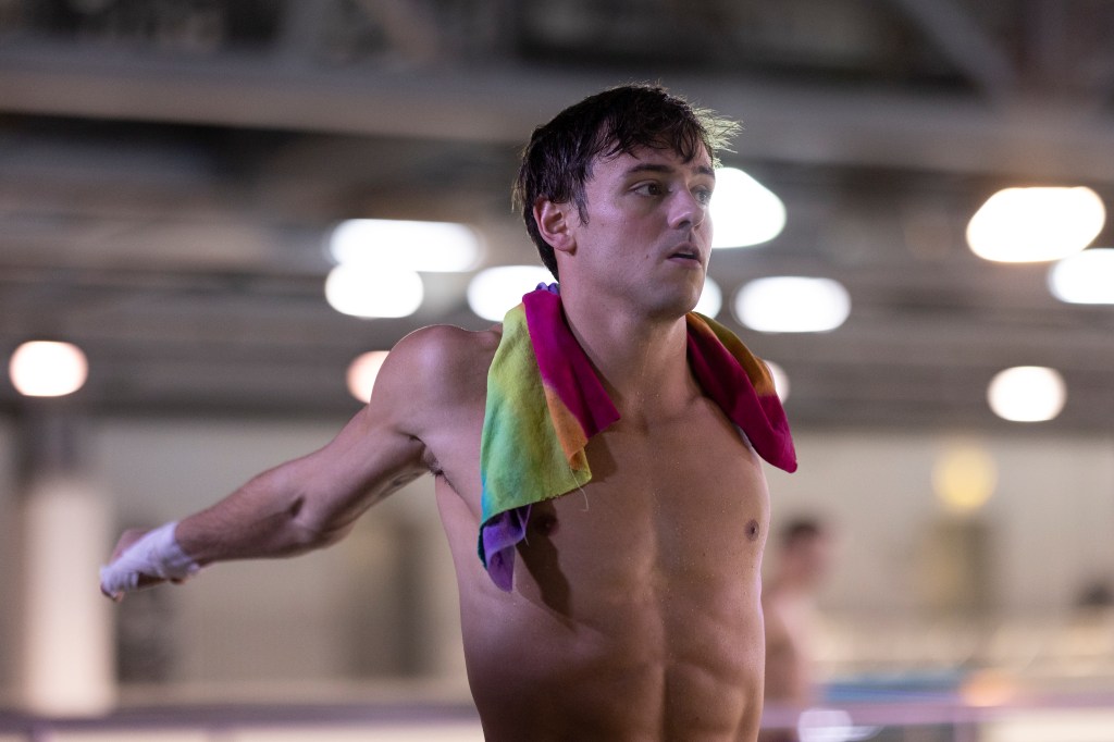 Tom Daley stretching his arms behind his back with a rainbow towel around his neck.