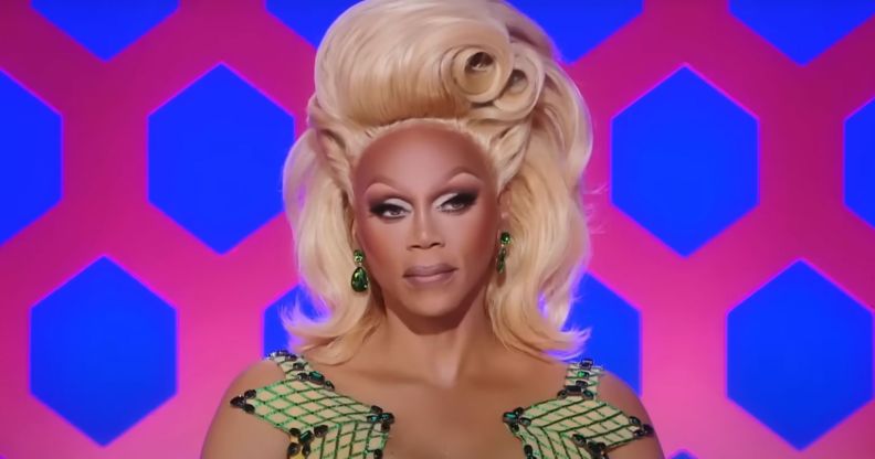 A still of RuPaul from RuPaul's Drag Race wearing a green patterned dress and blonde wig.