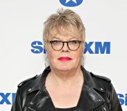Suzy Izzard wears black rimmed glasses, a black leather jacket, and hooped earrings as she poses on a red carpet.