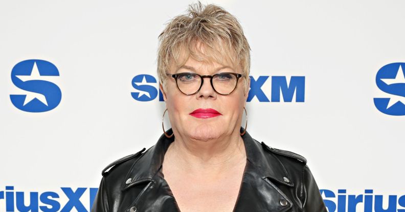 Suzy Izzard wears black rimmed glasses, a black leather jacket, and hooped earrings as she poses on a red carpet.